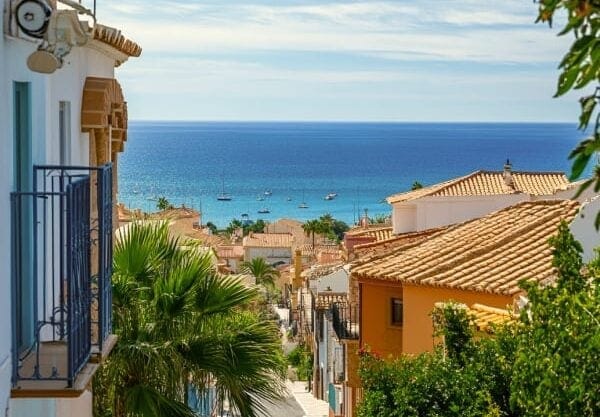 Charming Mediterranean street with colorful Spanish houses, sea views, and boats floating on calm blue waters.